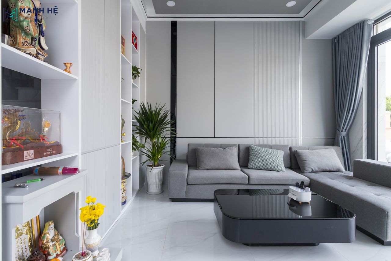 Bố trí ghế sofa chữ L với tone màu xám trung tính, kết hợp cùng bàn trà mặt kính đen tạo điểm nhấn cho không gian, bố trí thêm cây xanh sinh động