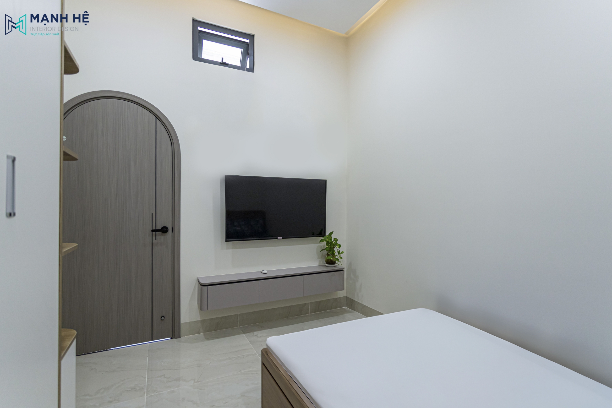 Kệ tivi treo tường màu trung tính, đối diện giường ngủ – điểm nhấn tiện nghi cho căn phòng nhỏ với cửa gỗ vòm độc đáo