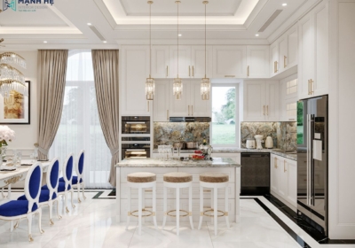 Không gian bếp biệt thự với hệ tủ bếp chữ L màu trắng, đảo bếp trung tâm mặt đá vân xanh, đèn thả pha lê và bàn ăn tân cổ điển kết hợp ghế nhung xanh dương nổi bật