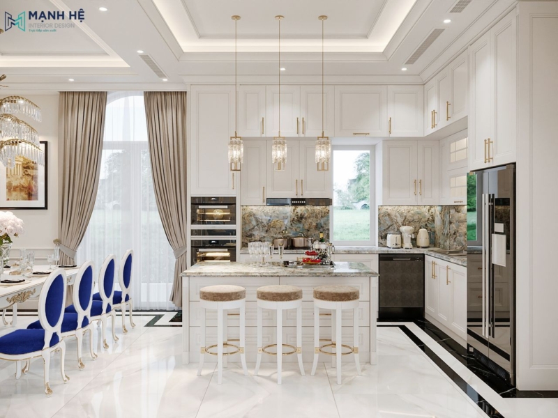 Không gian bếp biệt thự với hệ tủ bếp chữ L màu trắng, đảo bếp trung tâm mặt đá vân xanh, đèn thả pha lê và bàn ăn tân cổ điển kết hợp ghế nhung xanh dương nổi bật