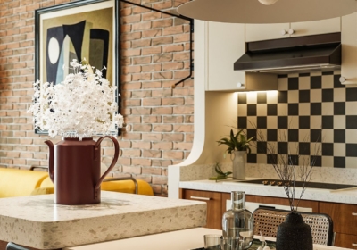 Cận cảnh đảo bếp liền bàn ăn phong cách Mid-century Modern, mặt đá terrazzo, ghế mây đệm xanh, nền tường gạch thô và ốp bếp họa tiết caro
